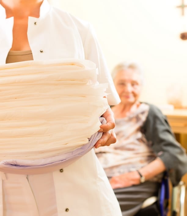 Woman carrying neatly folded laundry with a senior in the background, representing the return of clean, finished laundry that pickup and delivery service provides