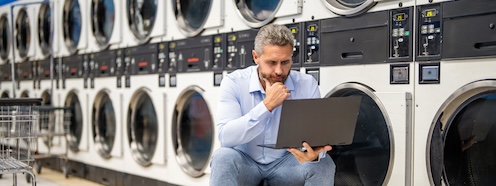 Man in business attire working on his laptop while seated near washing machines, representing the collision between a demanding professional schedule and the time laundry requires