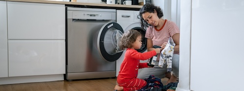 Parent showing a child an article of clothing while loading the washing machine together, representing how family laundry weaves into the constant demands of parenting