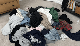 A large pile of unsorted laundry spread across a carpeted bedroom floor, featuring a mix of casual clothing including hoodies, jeans, t-shirts, flannel shirts, and socks in various colors — predominantly black, navy, gray, and blue — with a wooden dresser and bookshelf visible in the background.