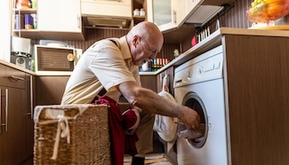 Older man bending down to reach a front-loading washing machine, illustrating the physical demands laundry places on seniors and the risk those demands quietly carry