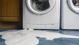 Water pooled on the floor in front of a washing machine, representing the sudden disruption of a broken appliance and the laundry problem it leaves behind
