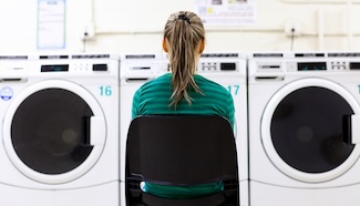 Person sitting alone in a laundromat facing a row of washing machines, capturing the tedium of waiting out another laundry cycle